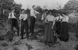 Men and women in a rural setting pose with rakes and hoes on a farming holiday in an English utopian community, probably around 1910.